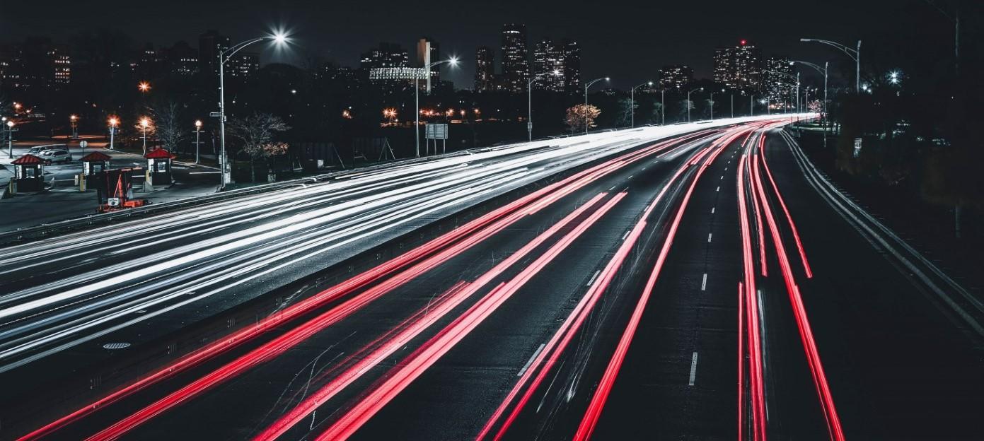 Long-exposure photograph of a highway at night showing red and white light trails from fast-moving vehicles, with a city skyline visible in the background.
